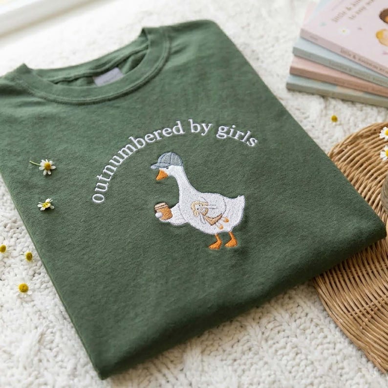 May include: Green sweatshirt with the embroidered phrase "outnumbered by girls" above a cartoon goose wearing a cap and holding a coffee cup. The goose has a rabbit design on its body. The sweatshirt is folded on a white surface.