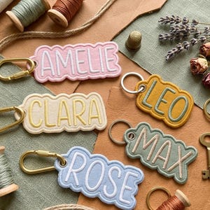 May include: Five embroidered fabric keychains with scalloped edges, each featuring a name in block lettering: AMELIE in pink, CLARA in yellow, LEO in mustard, MAX in sage green, and ROSE in light blue. Each keychain includes a metal clasp or ring.