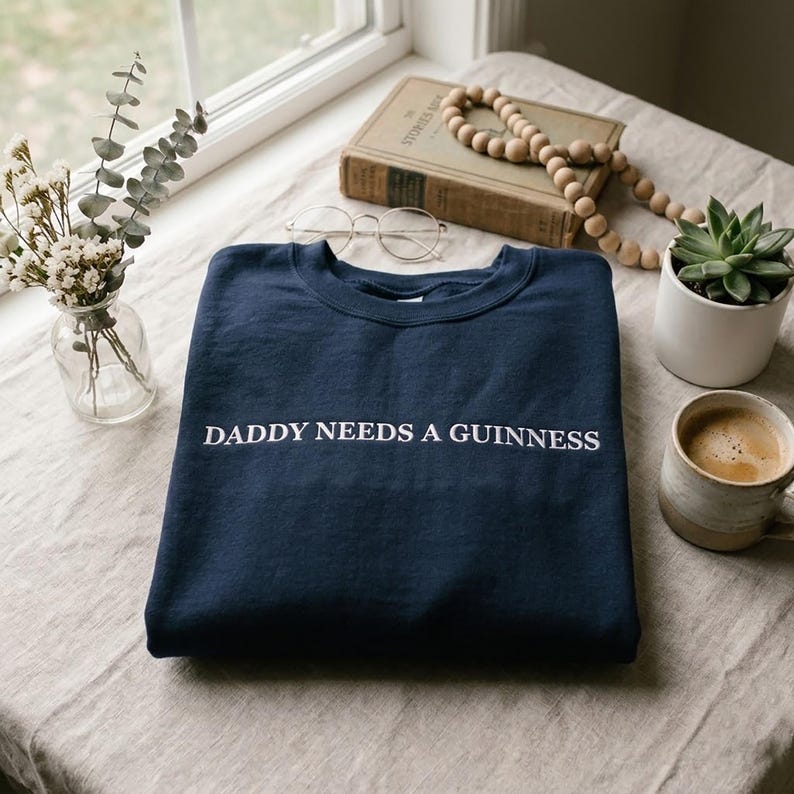 Op de afbeelding: Een marineblauw sweatshirt met de tekst "DADDY NEEDS A GUINNESS" in het wit. Het sweatshirt ligt opgevouwen op een tafel naast een kop koffie, een vetplant en een vintage boek.