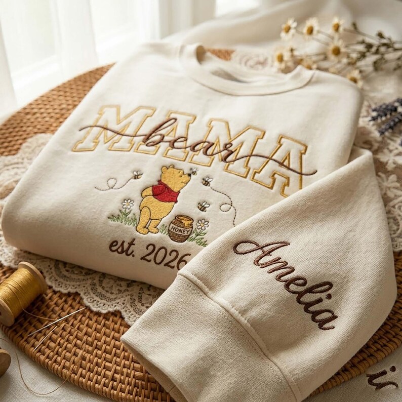 May include: Cream-colored sweatshirt with embroidered "Mama Bear" text in gold and brown. Features a Winnie the Pooh design with bees, flowers, and "est. 2026". The sleeve is embroidered with the name "Amelia".