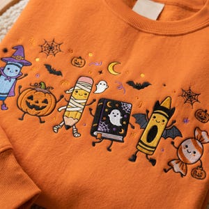 May include: An orange crewneck sweatshirt featuring colorful embroidered Halloween-themed characters including a crayon dressed as a witch, a mummy pencil, a ghost, a book, a bat-winged crayon, and a candy piece, surrounded by small pumpkins and webs.