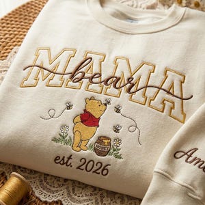 May include: Cream-colored sweatshirt with embroidered "MAMA bear" text and a Winnie the Pooh design. The design includes Pooh Bear, bees, flowers, a honey pot, and the text "est. 2026".
