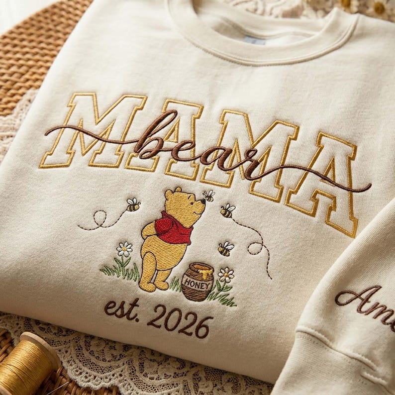May include: Cream-colored sweatshirt with embroidered "MAMA bear" text and a Winnie the Pooh design. The design includes Pooh Bear, bees, flowers, a honey pot, and the text "est. 2026".