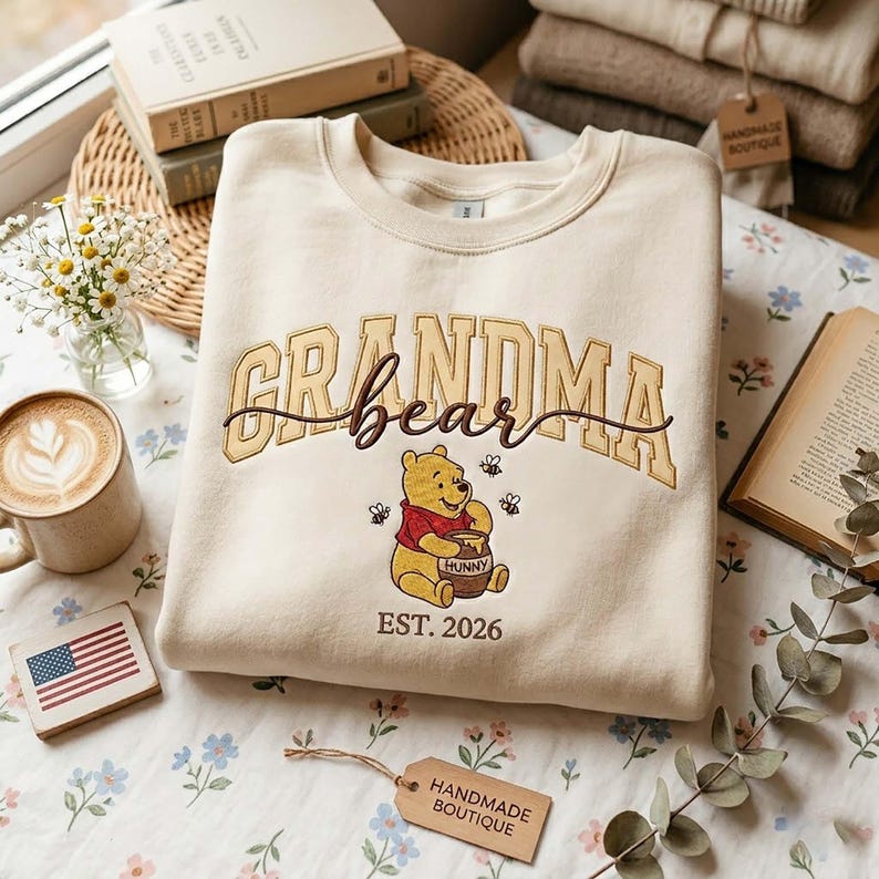May include: Cream-colored sweatshirt with the words "GRANDMA bear" in a gold arched font and a brown script font. A Winnie the Pooh graphic is below the text, holding a honey pot. The text "EST. 2026" is below the graphic.