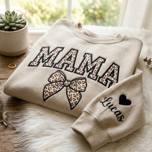 Pode incluir: Camisola bege com a palavra "MAMA" bordada em letras grandes com estampa de leopardo. Uma fita com estampa de leopardo está abaixo da palavra. A manga tem o nome "Lucas" e um coração preto.