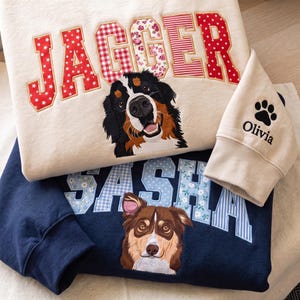 Peut inclure: Un sweat-shirt crème avec le nom JAGGER en lettres de tissu à motifs rouges et un bouvier bernois brodé, avec une manche portant le nom Olivia et une empreinte de patte. En dessous, un sweat-shirt bleu marine affiche SASHA en lettres à motifs bleus.