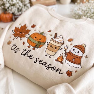 Op de afbeelding: Een crèmekleurig sweatshirt met een geborduurd ontwerp van een lachend esdoornblad, een pompoen met een groene sjaal, een koffiebeker met slagroom en een spookje met een bruine muts en trui, met de tekst 'tis the season'.