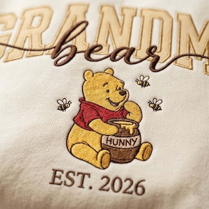 May include: Cream-colored sweatshirt with embroidered text and a cartoon bear. The text reads "Grandma Bear" in tan and brown thread, with "EST. 2026" below. The bear is yellow, wearing a red shirt, and holding a honey pot.