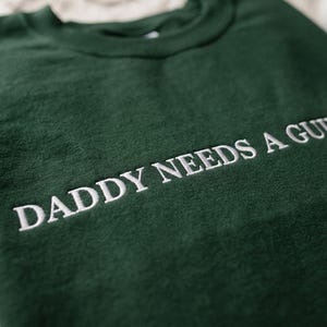 Op de afbeelding: Een donkergroene sweatshirt met de geborduurde tekst "DADDY NEEDS A GUN". De witte tekst contrasteert met de donkergroene stof. De sweatshirt heeft een ronde hals en een relaxte pasvorm, ideaal voor casual wear.