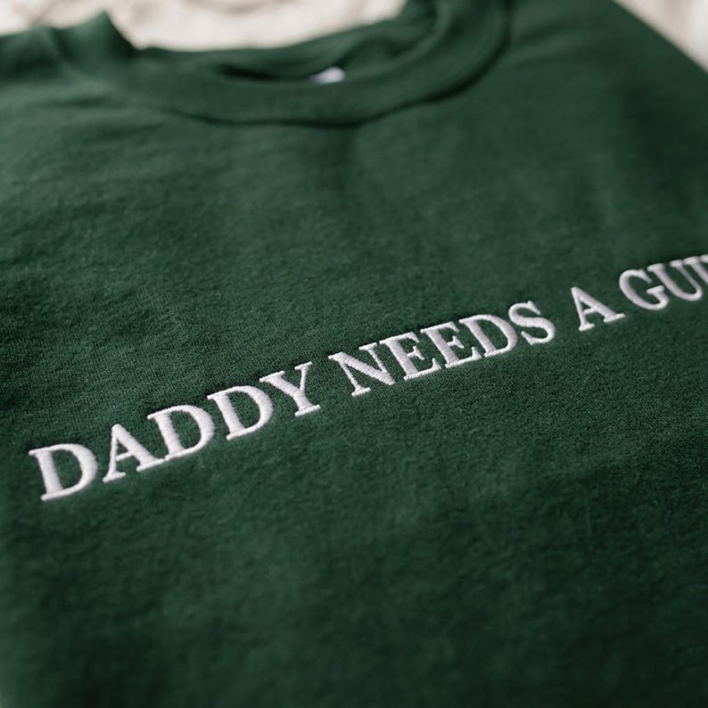Op de afbeelding: Een donkergroene sweatshirt met de geborduurde tekst "DADDY NEEDS A GUN". De witte tekst contrasteert met de donkergroene stof. De sweatshirt heeft een ronde hals en een relaxte pasvorm, ideaal voor casual wear.