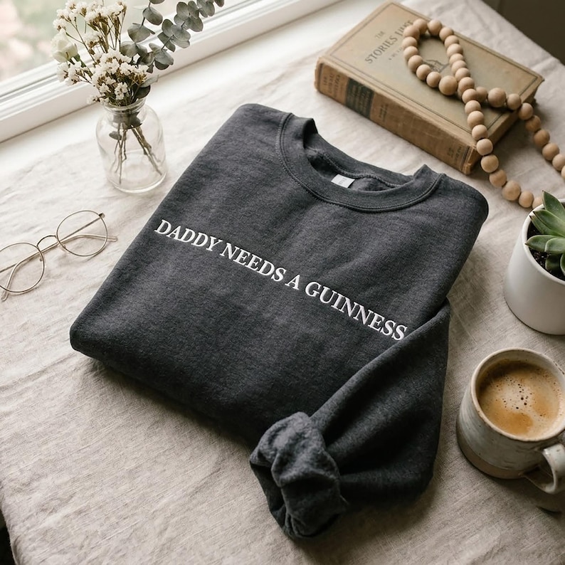 Op de afbeelding: Donkergrijze sweatshirt met de witte tekst "DADDY NEEDS A GUINNESS". De opgevouwen sweatshirt ligt op een neutraal gekleurd oppervlak, samen met een kopje koffie, een boek en een kleine plant.