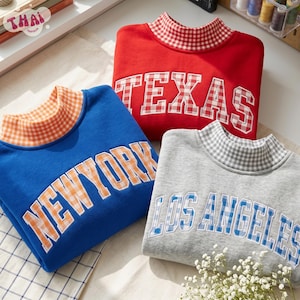 May include: Three mock neck sweatshirts featuring city names in block letters. A red sweatshirt with TEXAS in red gingham, a blue sweatshirt with NEW YORK in orange gingham, and a gray sweatshirt with LOS ANGELES in blue gingham, all with matching collars.