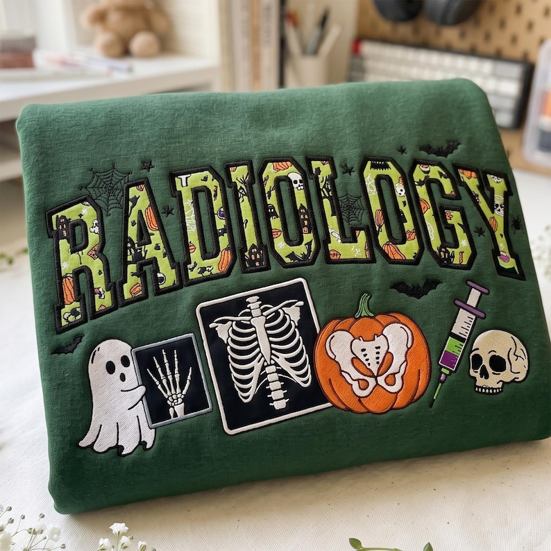 May include: A dark green sweatshirt featuring the embroidered word RADIOLOGY in a varsity font with a Halloween pattern. Below, there is an embroidered ghost holding an X-ray of a hand, a rib cage X-ray, a pumpkin with a pelvis, a syringe, and a skull.