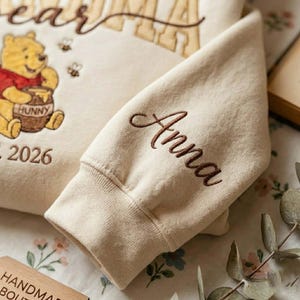 May include: Cream-colored sweatshirt featuring an embroidered Winnie the Pooh graphic with the word "Hunny" on the honey pot. The sleeve is embroidered with the name "Anna" in brown script. The year "2026" is also embroidered on the sweatshirt.