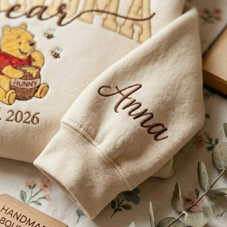 May include: Cream-colored sweatshirt featuring an embroidered Winnie the Pooh graphic with the word "Hunny" on the honey pot. The sleeve is embroidered with the name "Anna" in brown script. The year "2026" is also embroidered on the sweatshirt.