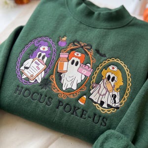 May include: A dark green sweatshirt featuring embroidered designs of three ghosts dressed as nurses in ornate frames. The ghosts hold medical items like a clipboard, a pill bottle, and a syringe. The text HOCUS POKE-US is embroidered below.
