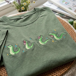 May include: Olive green t-shirt featuring an embroidered design of four green cartoon dinosaurs with open mouths and star accents. The t-shirt is made of a soft material and is folded on a woven surface.