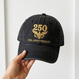 May include: Black baseball cap with gold embroidered text and design. The cap features the number "250" above an eagle emblem, with "USA ANNIVERSARY" below. The cap is made of a dark, possibly denim-like material.