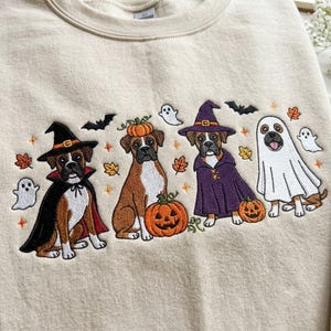 May include: A beige sweatshirt featuring an embroidered design of four boxer dogs in Halloween costumes. The dogs are depicted as a vampire, a pumpkin-topped dog, a witch, and a ghost, surrounded by small embroidered ghosts, bats, and leaves.