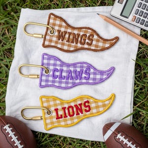 May include: Three pennant-shaped keychains with gold-toned metal rings. The top keychain is brown gingham with the text WINGS, the middle is purple gingham with the text CLAWS, and the bottom is yellow gingham with the text LIONS in red embroidery.