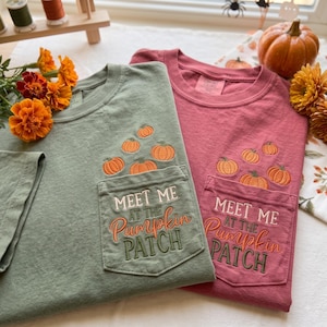May include: Two short-sleeved t-shirts in sage green and dusty rose, each featuring a chest pocket embroidered with orange pumpkins and the text "MEET ME AT THE PUMPKIN PATCH" in white, orange, and dark green thread, displayed on a flat surface.