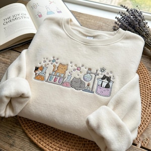Peut inclure: Sweat-shirt crème avec un motif brodé de chats de dessins animés et de matériel de laboratoire de chimie. Le motif comprend des béchers, des tubes à essai et des structures moléculaires. Le texte "THE JOY OF CHEMISTRY" est visible sur un livre ouvert en arrière-plan.