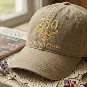 May include: A khaki baseball cap with the text "250 USA ANNIVERSARY" embroidered in gold. The design includes a golden eagle emblem. The cap is sitting on a table next to a magazine and a small American flag.