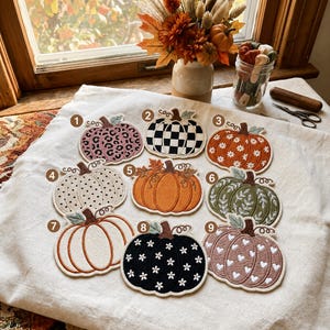 May include: Nine embroidered pumpkin patches arranged on a white fabric surface, each numbered one through nine. Designs include leopard print, black and white checkerboard, floral patterns, polka dots, solid orange, leaves, and white hearts.