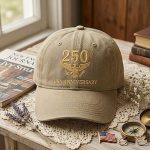 May include: A tan baseball cap with the text "250 USA ANNIVERSARY" embroidered in gold. The design includes a gold eagle emblem. The cap is sitting on a wooden surface with a book and other decorative items.