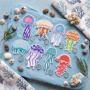May include: A collection of eight die-cut stickers featuring colorful jellyfish illustrations, each labeled with a small numbered glass token from one to eight. The stickers display various patterns and hues, including purple, orange, blue, and red.