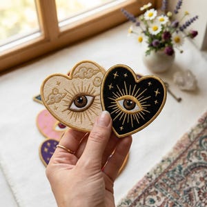 May include: Two heart-shaped embroidered patches featuring a central eye design with radiating sunburst lines. One patch is cream-colored with cloud motifs, while the other is black with star accents. Both patches have gold-colored stitched borders.