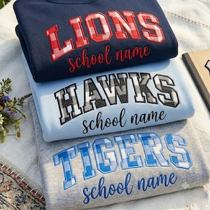 Op de afbeelding: Drie gevouwen sweaters met ronde hals in marineblauw, lichtblauw en heidegrijs. Elk heeft geborduurde letters in varsity-stijl met rode, zwarte en blauwe ruitpatronen, met de woorden LIONS, HAWKS en TIGERS boven de tekst school name.