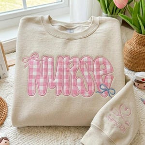 May include: A cream-coloured sweatshirt embroidered with the word "NURSE" in pink gingham. A blue bow is embroidered below the "E". The sleeve of a matching sweatshirt features a stethoscope and heart design with the text "Avery, RN".