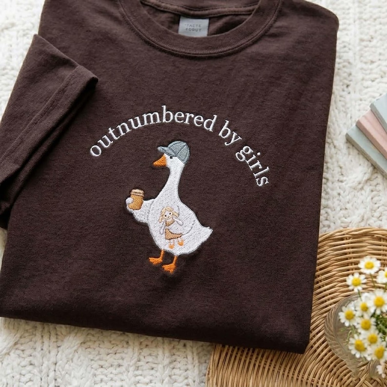 May include: A dark brown t-shirt with the embroidered phrase "outnumbered by girls." The shirt features a cartoon goose wearing a hat and holding a cup, with a smaller character on its chest. The shirt is made of a soft material.
