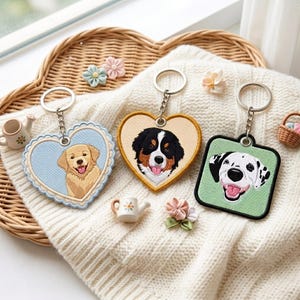 May include: Three keychains featuring embroidered dog portraits: a golden retriever on a light blue heart, a Bernese mountain dog on a yellow heart, and a Dalmatian on a green square. Each has a metal ring and chain attached to the top.