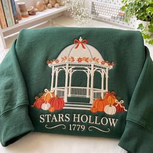 May include: A dark green sweatshirt featuring an embroidered white gazebo topped with a red bow. The gazebo is surrounded by orange, red, and white pumpkins and autumn leaves. The text "STARS HOLLOW 1779" is embroidered in white below the gazebo.