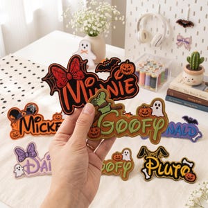 Op de afbeelding: Een collectie geborduurde stoffen patches met Disney-karakternamen in Halloween-thema's. De patches bevatten Minnie, Mickey, Goofy, Donald, Daisy en Pluto, versierd met pompoenen, spoken, vleermuizen en spinnenwebben.