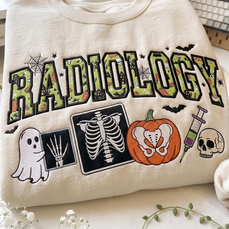 May include: A cream-colored sweatshirt featuring the word RADIOLOGY in large green letters with a Halloween pattern. Below the text are embroidered designs of a ghost holding an X-ray, a rib cage X-ray, an orange pumpkin, a syringe, and a skull.