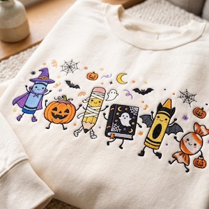 May include: A cream-colored crewneck sweatshirt featuring colorful embroidered Halloween-themed characters including a crayon dressed as a wizard, a jack-o'-lantern, a mummy pencil, a ghost book, a bat-winged crayon, and a candy piece.