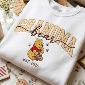 May include: White sweatshirt featuring an embroidered design. The design includes the words "Grandma Bear" in tan and brown, with a cartoon bear holding a honey pot. The text "EST. 2026" is below the bear.