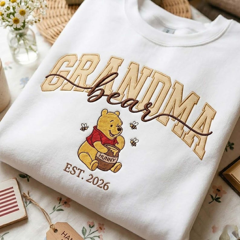 May include: White sweatshirt featuring an embroidered design. The design includes the words "Grandma Bear" in tan and brown, with a cartoon bear holding a honey pot. The text "EST. 2026" is below the bear.
