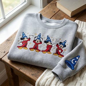 Pode incluir: Um moletom cinza com um desenho bordado do Mickey Mouse em traje de mago. O desenho inclui Mickey em uma túnica vermelha e chapéu de mago azul com estrelas. A manga tem um patch de chapéu de mago azul correspondente.