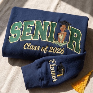 Op de afbeelding: Een marineblauw sweatshirt met groen en goudkleurig borduurwerk dat SENIOR spelt, waarbij de letter O is vervangen door een figuur met een afstudeerhoed. Daaronder staat Class of 2026 in goud. De mouw is voorzien van de naam Eleanor.