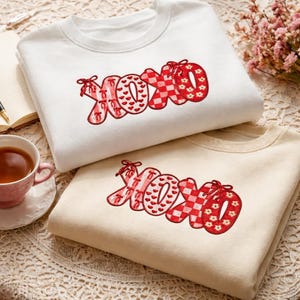 Może przedstawiać: Dwie bluzy, jedna biała i jedna beżowa, każda z haftem w postaci słowa "XOXO" w kolorze czerwonym. Litery mają różne wzory, w tym serca, kratę vichy i kwiatowe wzory. Bluzy są złożone i prezentowane na wzorzystej powierzchni.