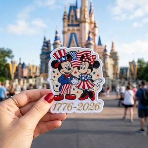 May include: A white embroidered patch featuring Mickey and Minnie Mouse dressed in patriotic red, white, and blue clothing with stars and stripes. The design includes an American flag and the gold text 1776-2026 on a cream-colored background.