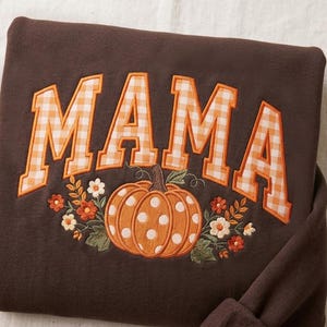 May include: A dark brown sweatshirt featuring the word MAMA in large, orange and white gingham block letters. Below the text is an embroidered orange pumpkin with white polka dots, surrounded by small orange and white flowers.