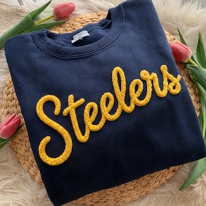 May include: Navy blue sweatshirt with the word "Steelers" in yellow, textured lettering. The crew neck sweatshirt is displayed on a woven surface with pink tulips and greenery.