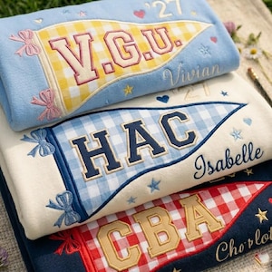 Könnte beinhalten: Drei gefaltete Sweatshirts mit gestickten Wimpelmotiven. Das oberste ist hellblau mit einem gelb karierten Wimpel und 'V.G.U.'-Text. Das mittlere ist cremefarben mit einem blau karierten Wimpel und 'HAC'. Das untere ist marineblau mit einem rot karierten Wimpel und 'CBA'.