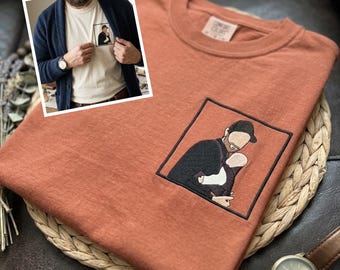 May include: Rust-colored t-shirt with an embroidered image of a person holding a baby. The image is framed in a black square. A small image of a person wearing the shirt is in the upper left corner. A watch with a brown leather band is in the lower right corner.