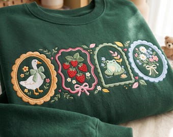 May include: A dark green crewneck sweatshirt featuring four embroidered rectangular and oval frames. The designs include a white goose with a green bow, a cluster of red strawberries, a green frog on a lily pad, and a bouquet of colorful flowers.
